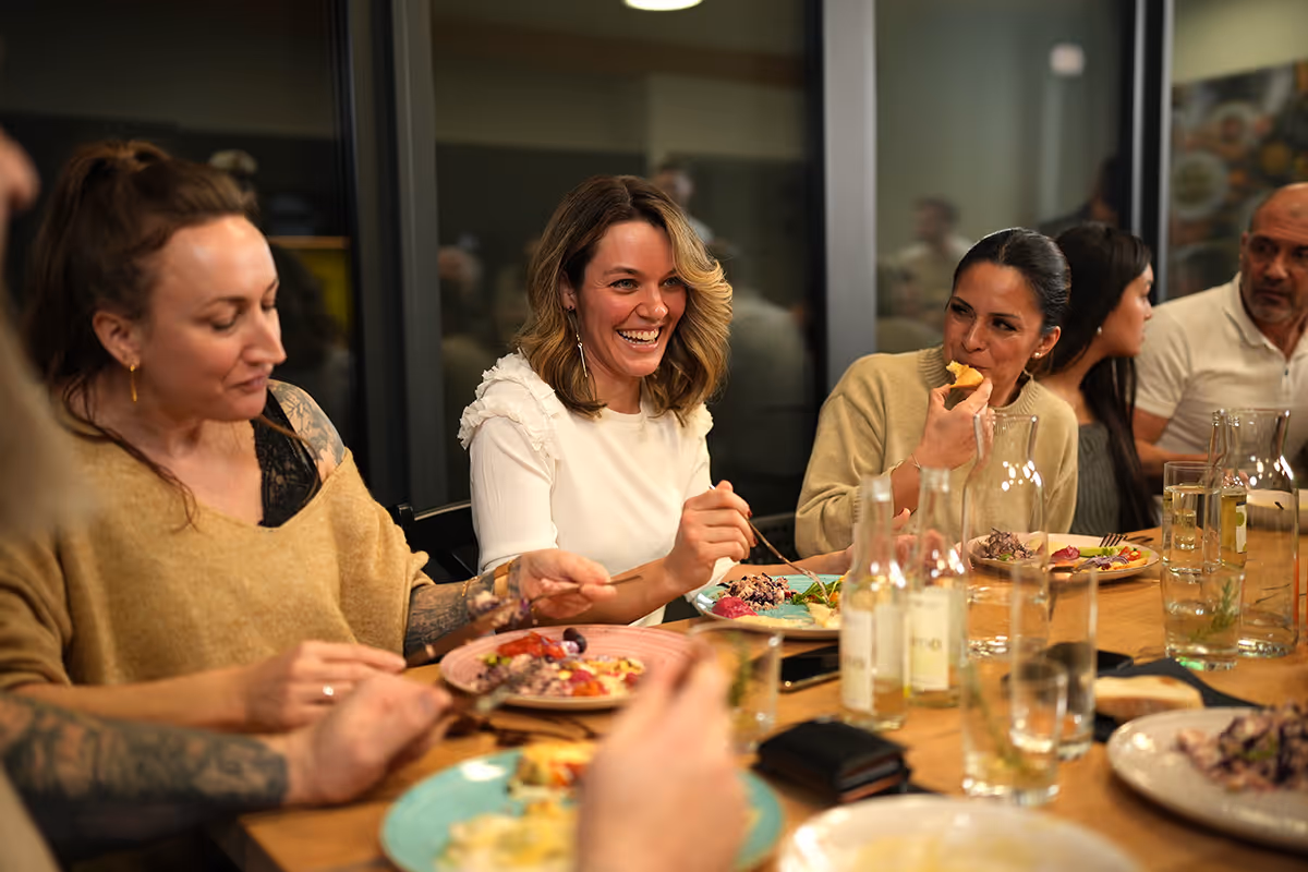 Eine Gruppe freudig lachender Kursteilnehmer beim gemeinsamen Essen am Tisch.